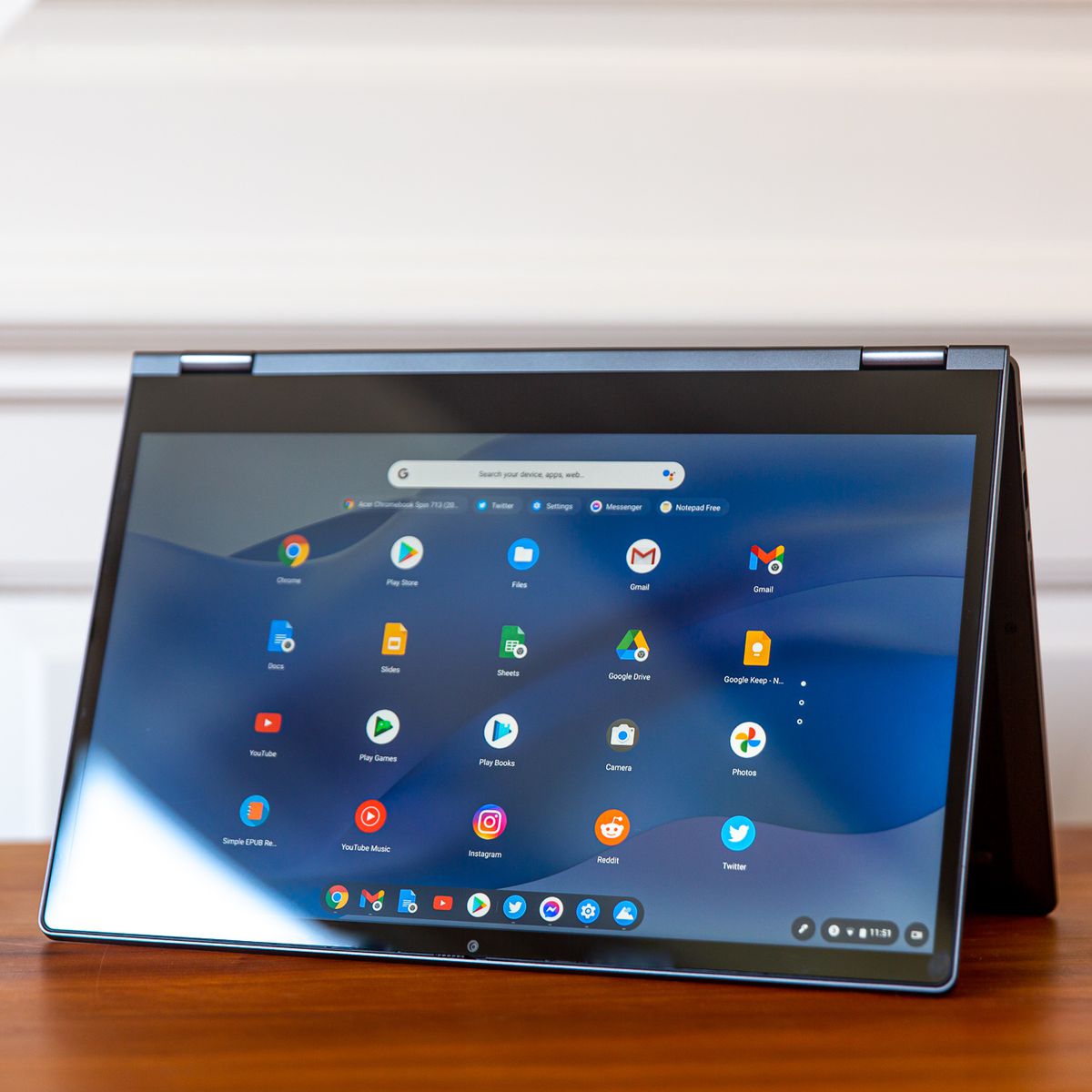 Chromebook Lenovo Flex 5 w trybie namiotu, pochylony w lewo.  Ekran wyświetla siatkę ikon systemu operacyjnego Chrome na niebieskim, falistym tle.