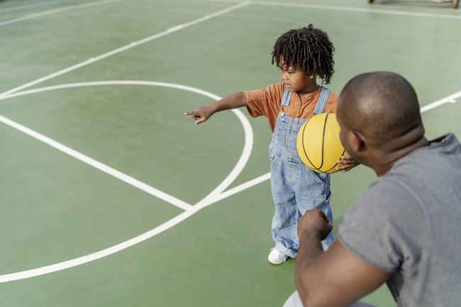 Czarne dziecko trzyma w dłoni żółtą piłkę do koszykówki i wskazuje boisko sportowe.