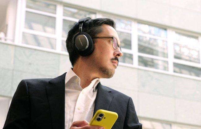 Redaktor Giga, Stefan Bubeck, nosi czarne słuchawki nad-douszne Bowers & Wilkins PX7 S3 i trzyma żółty smartfon. W tle fasada nowoczesnego budynku można zobaczyć z dużymi oknami.